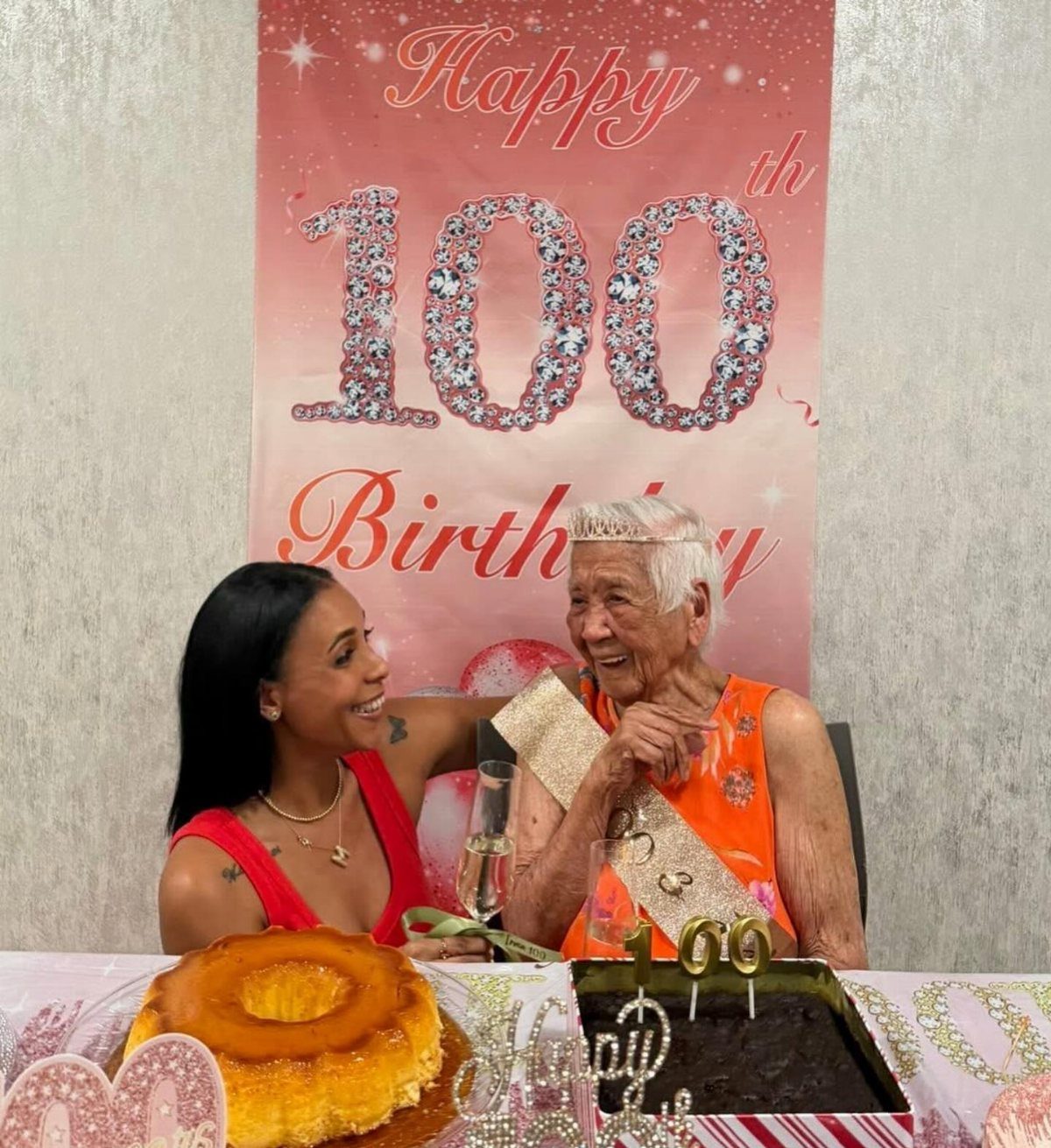 An elderly woman and a younger woman smiling together at a 100th birthday celebration table.