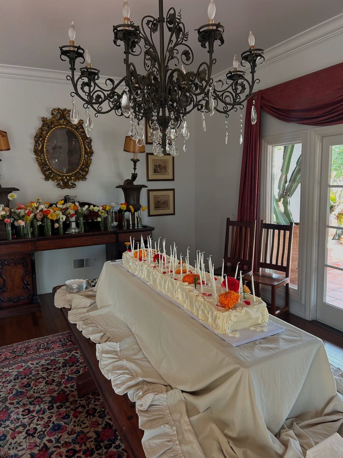 A long white frosted cake decorated with many thin candles and bright flowers on a draped table.