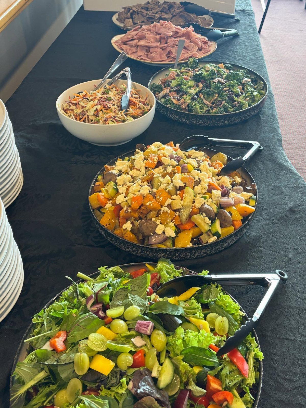 Various large salad bowls and meat platters arranged on a long black banquet table.