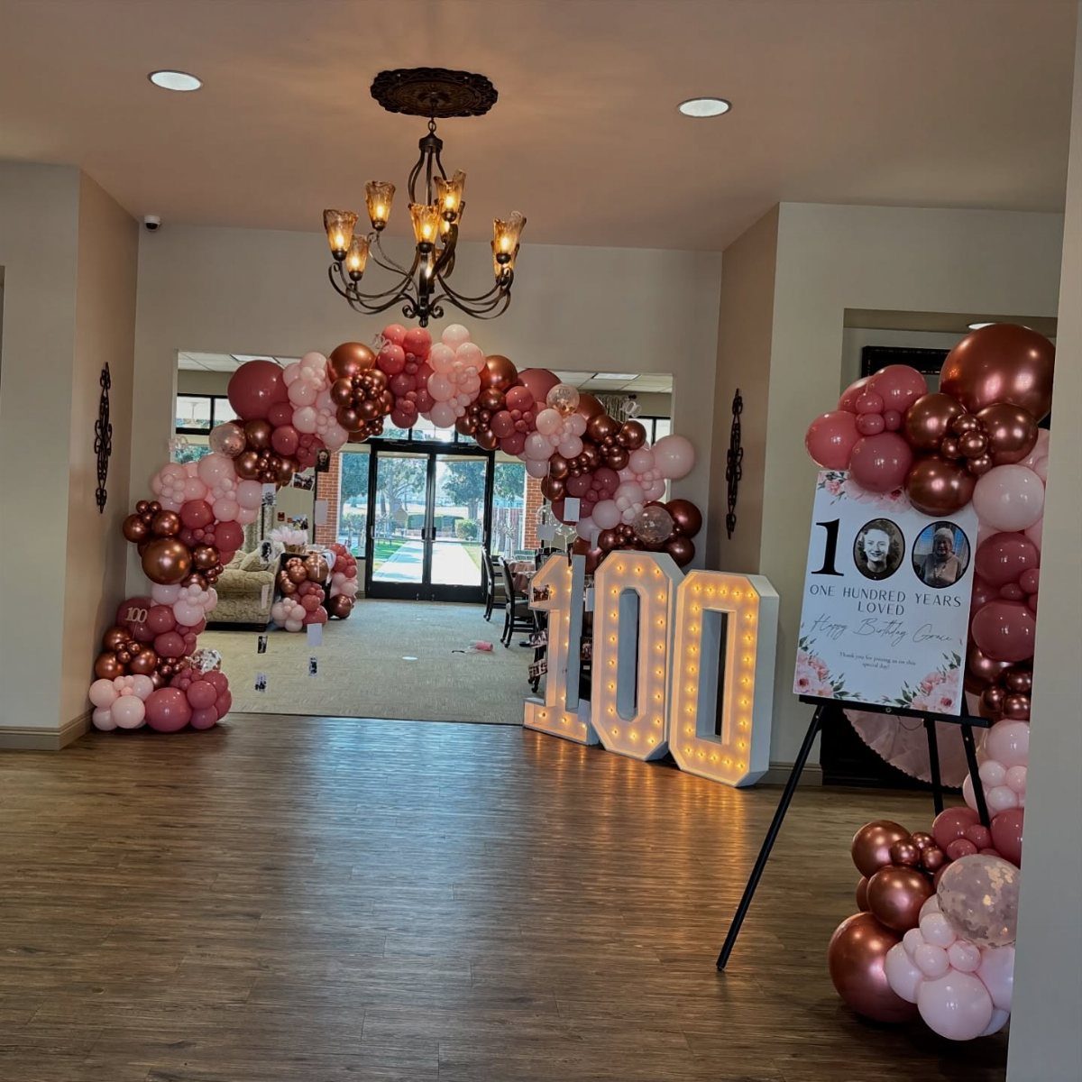 Pink and rose gold balloon arch surrounding large marquee numbers spelling 100 in a decorated room.