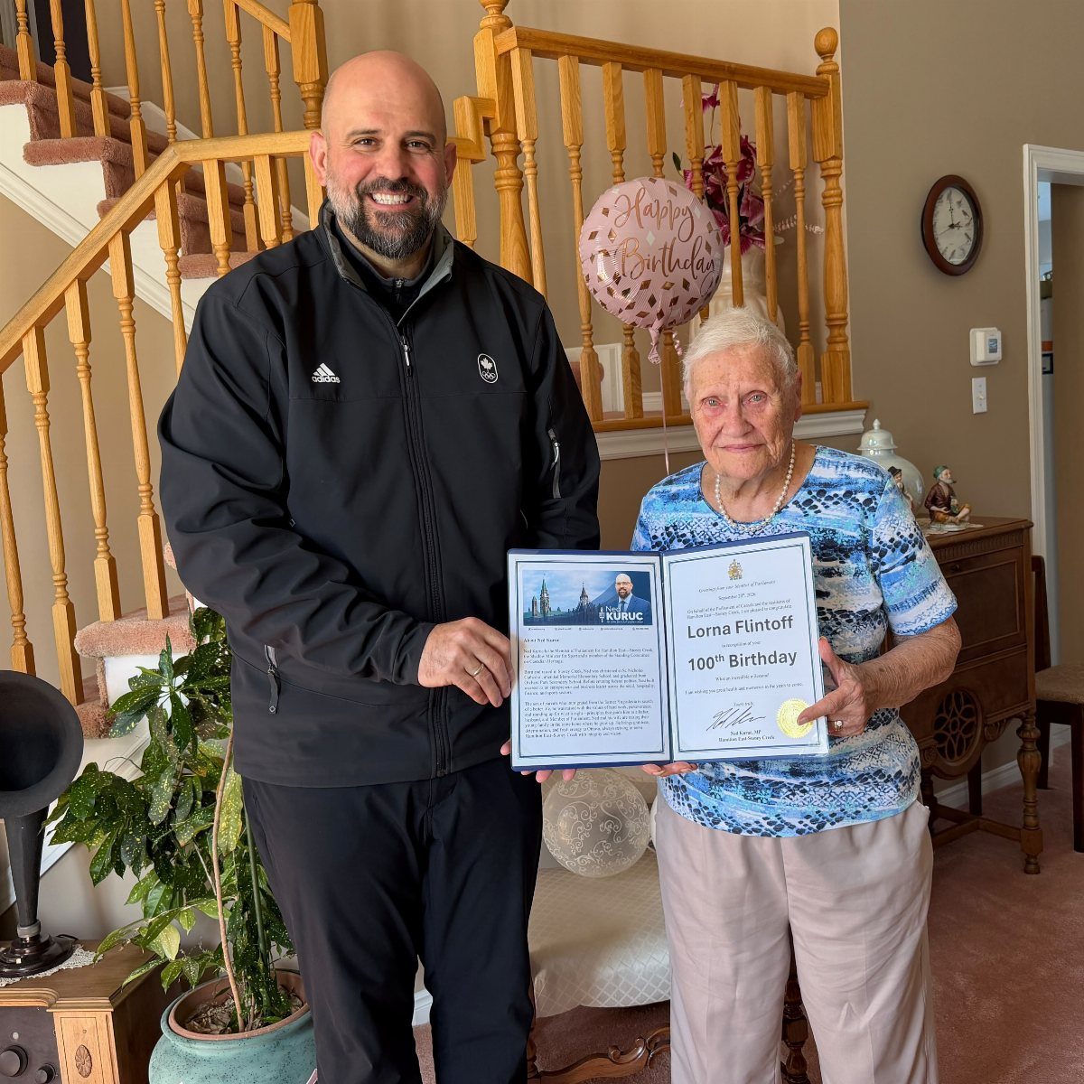 A man and an elderly woman holding a formal 100th birthday recognition certificate.