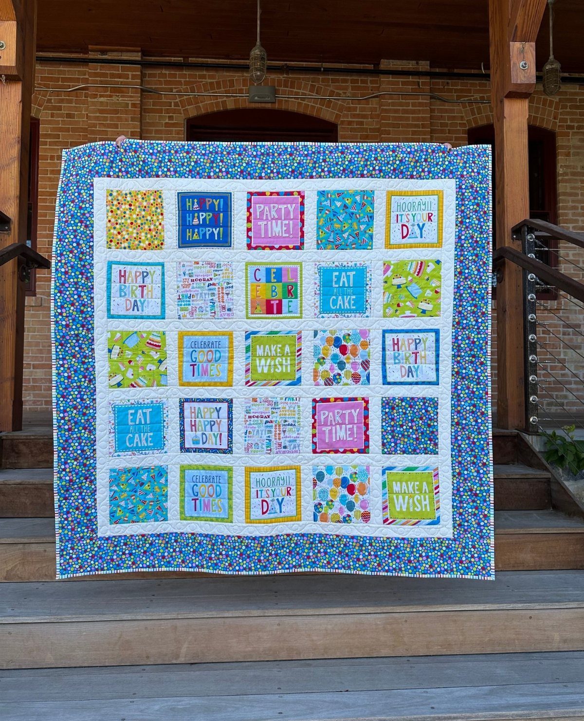 A colorful patchwork quilt with celebratory phrases hanging on wooden outdoor stairs.