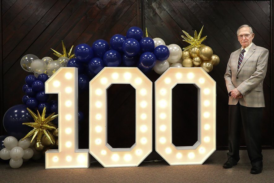 Elderly man standing beside large illuminated marquee numbers spelling one hundred with blue and gold balloons.