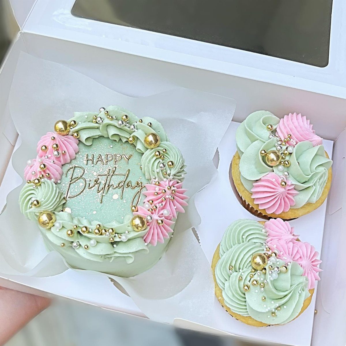 Small green and pink frosted cake and two cupcakes in a white box.