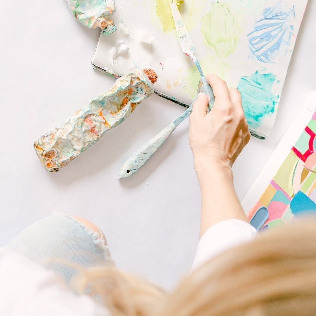 Hands using a palette knife to spread thick, colorful paint onto a canvas.