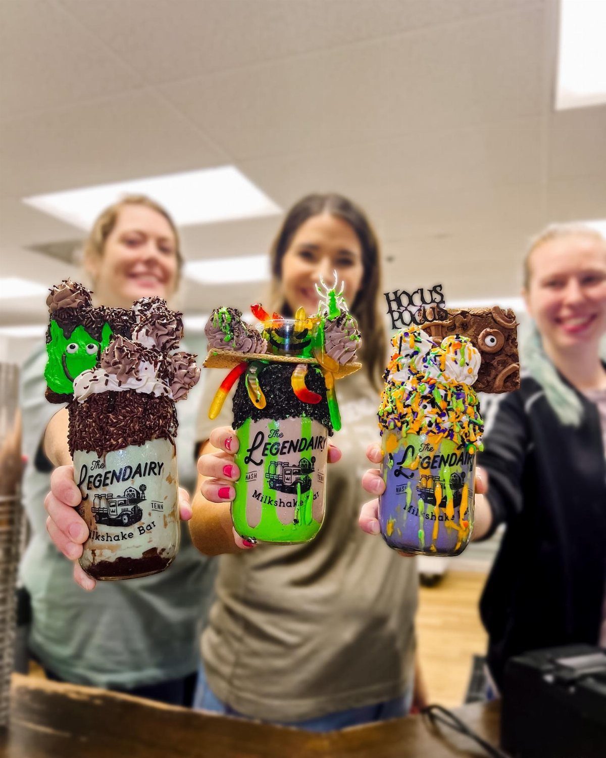 Three smiling women holding up large, highly decorated milkshakes with colorful toppings and sprinkles.