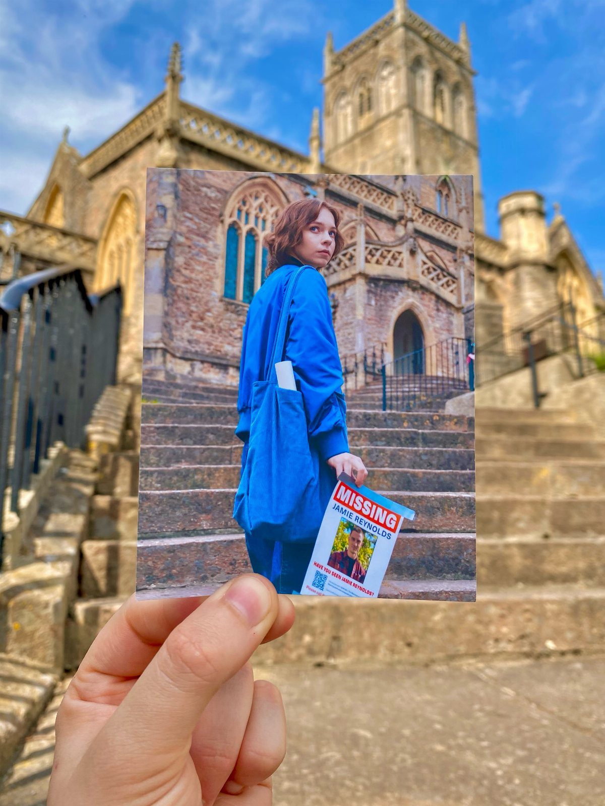 A hand holding a photograph of a woman with a missing person flyer against a cathedral.