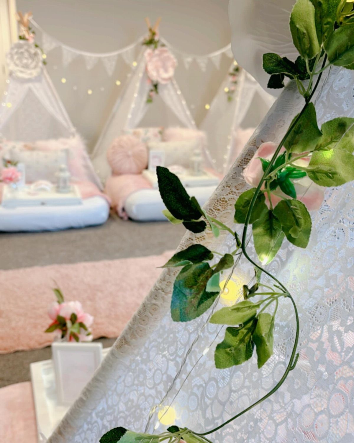 White lace teepee tents decorated with pink flowers and fairy lights on a pink rug.