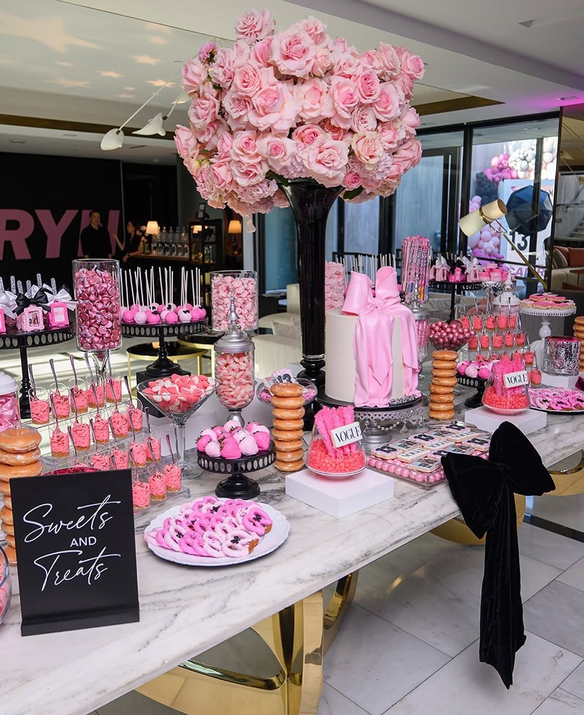 A dessert table featuring various pink candies, cupcakes, macarons, and a large bouquet of pink roses.