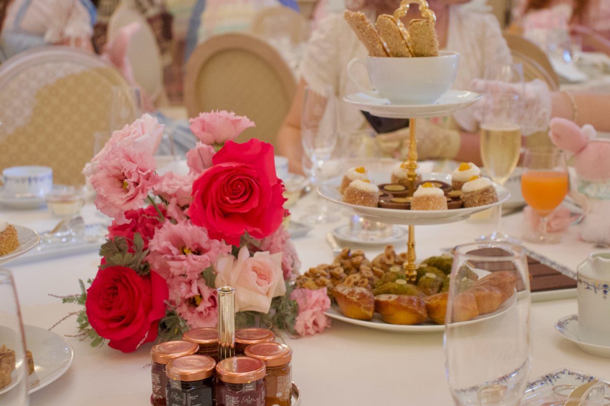 Pink roses and tiered dessert stands sit on a white tablecloth with various drinks and small jam jars.