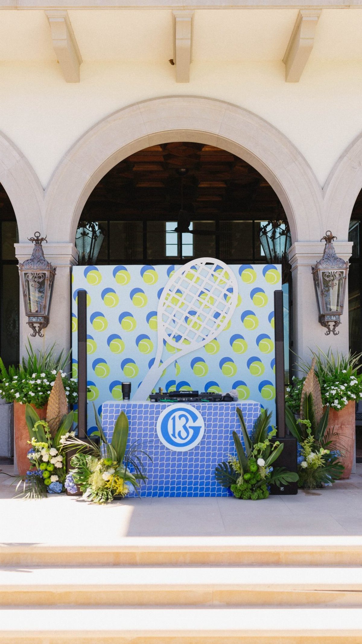 Tennis themed party setup with a blue mosaic table, large racket backdrop, and floral arrangements.