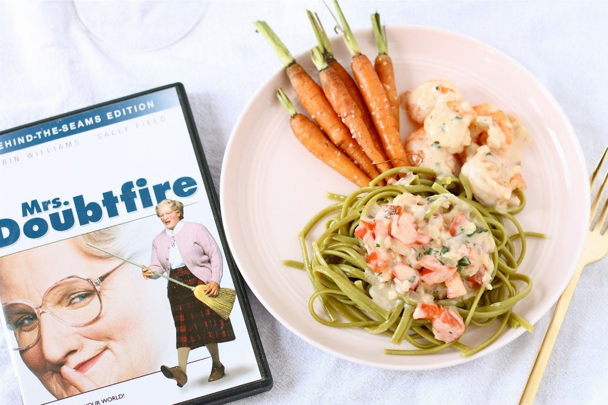 A DVD of Mrs. Doubtfire next to a plate of green pasta, shrimp, and carrots.