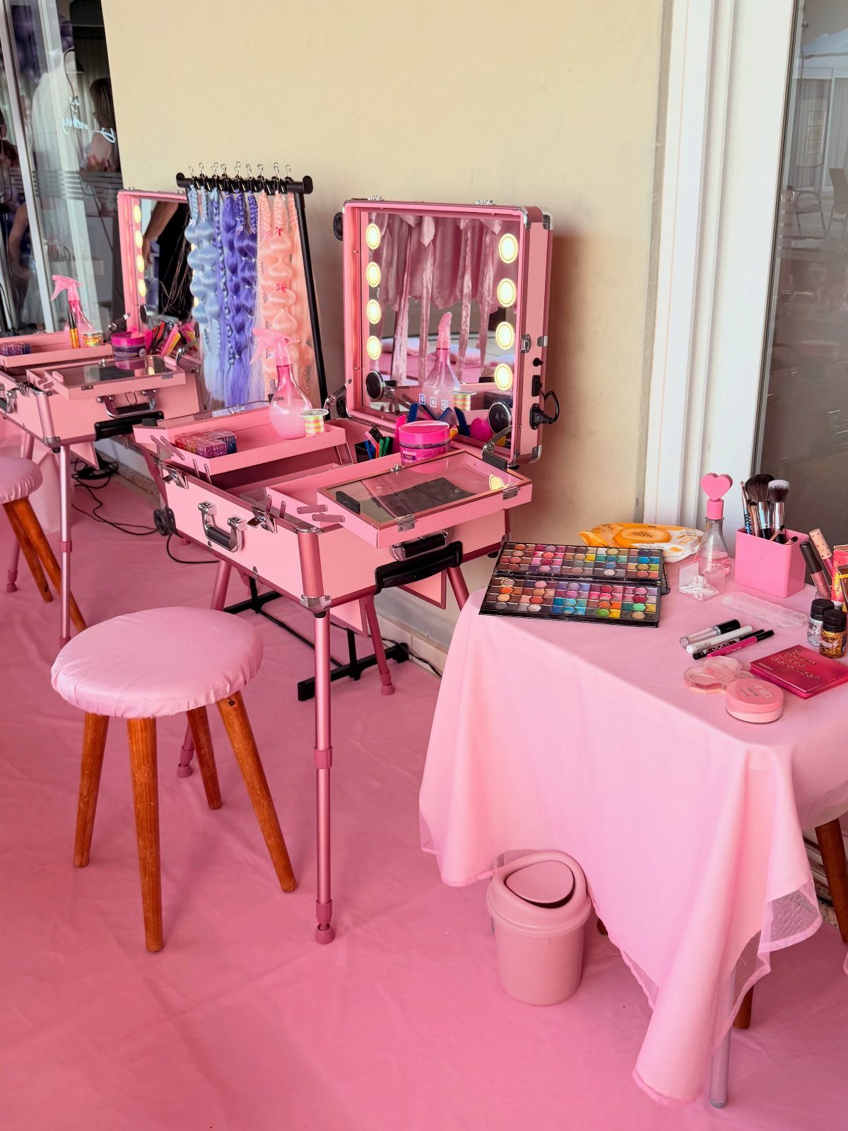 Pink makeup stations with lighted cases, cosmetics, and a pink stool on a pink floor.