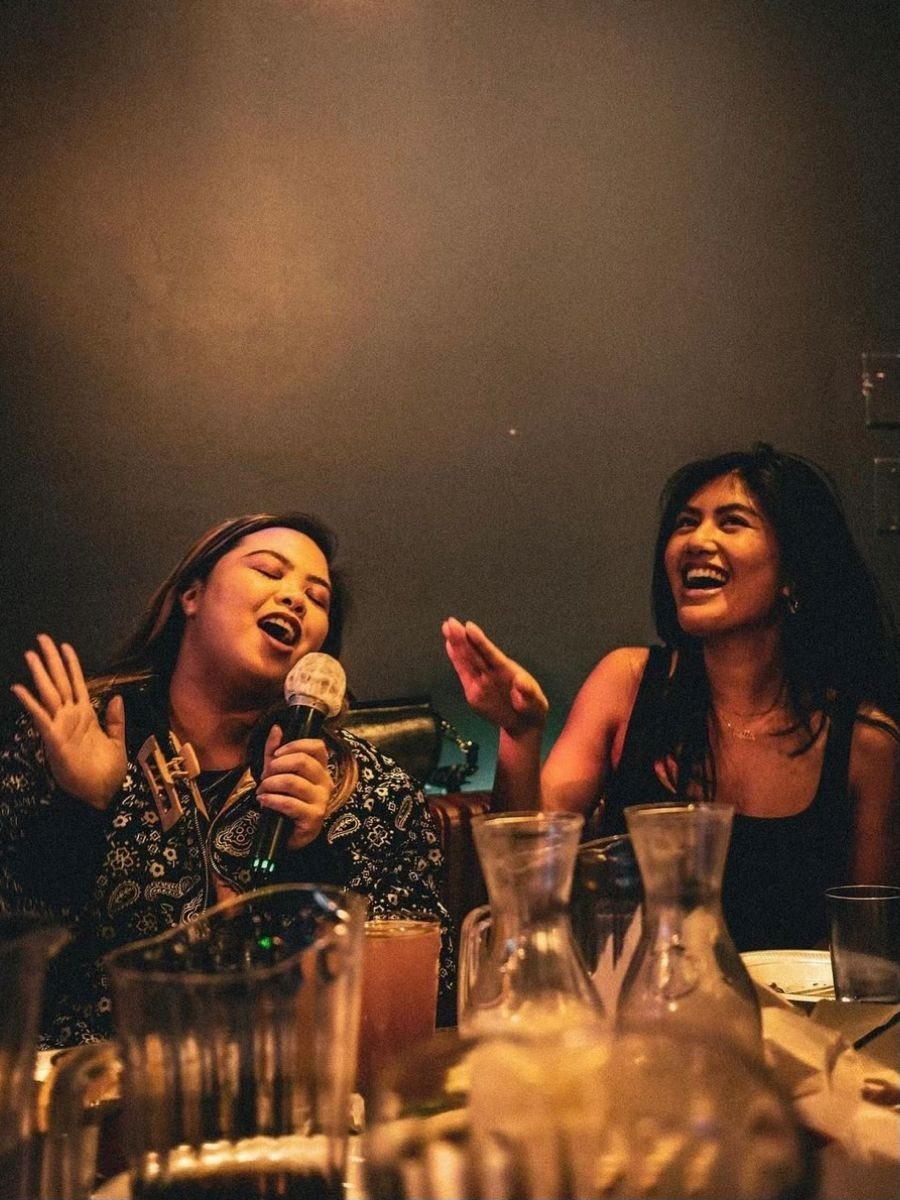 Two young women laughing and singing into a microphone at a dimly lit gathering.