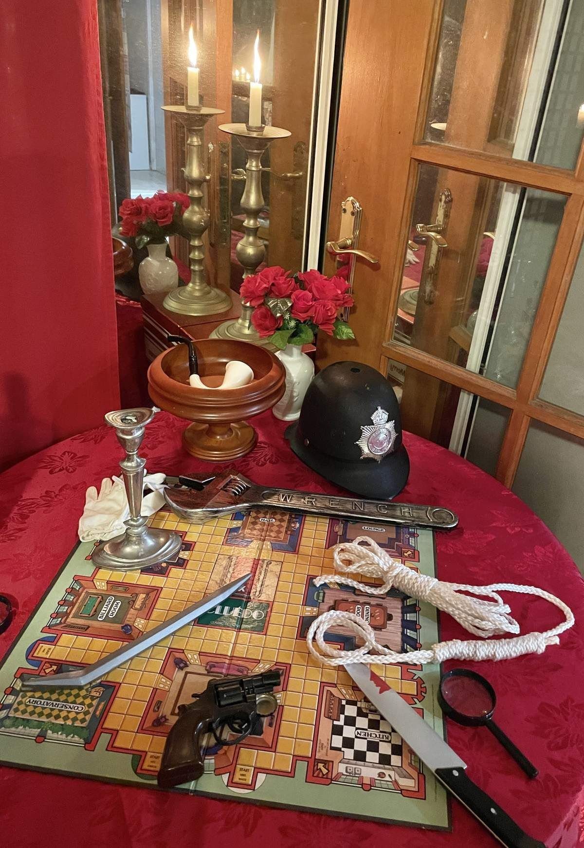 Mystery game props including a Clue board, revolver, knife, and police hat on a red table.
