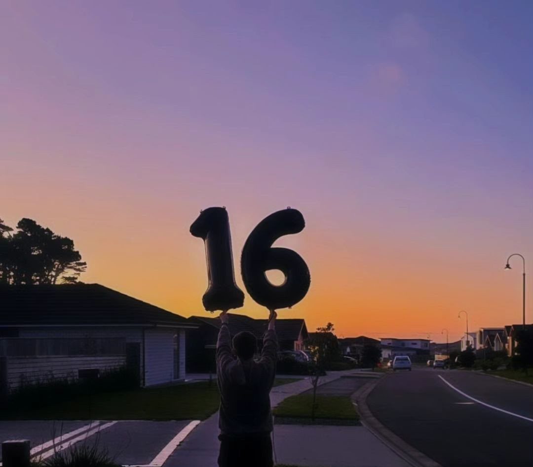 A person holding large number sixteen balloons against a colorful sunset sky in a residential area.