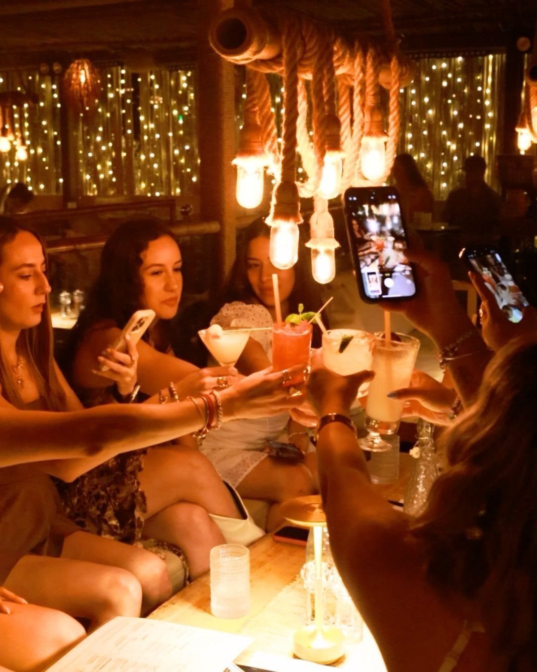 Young women clinking cocktail glasses together in a warm, dimly lit setting with glowing lights.