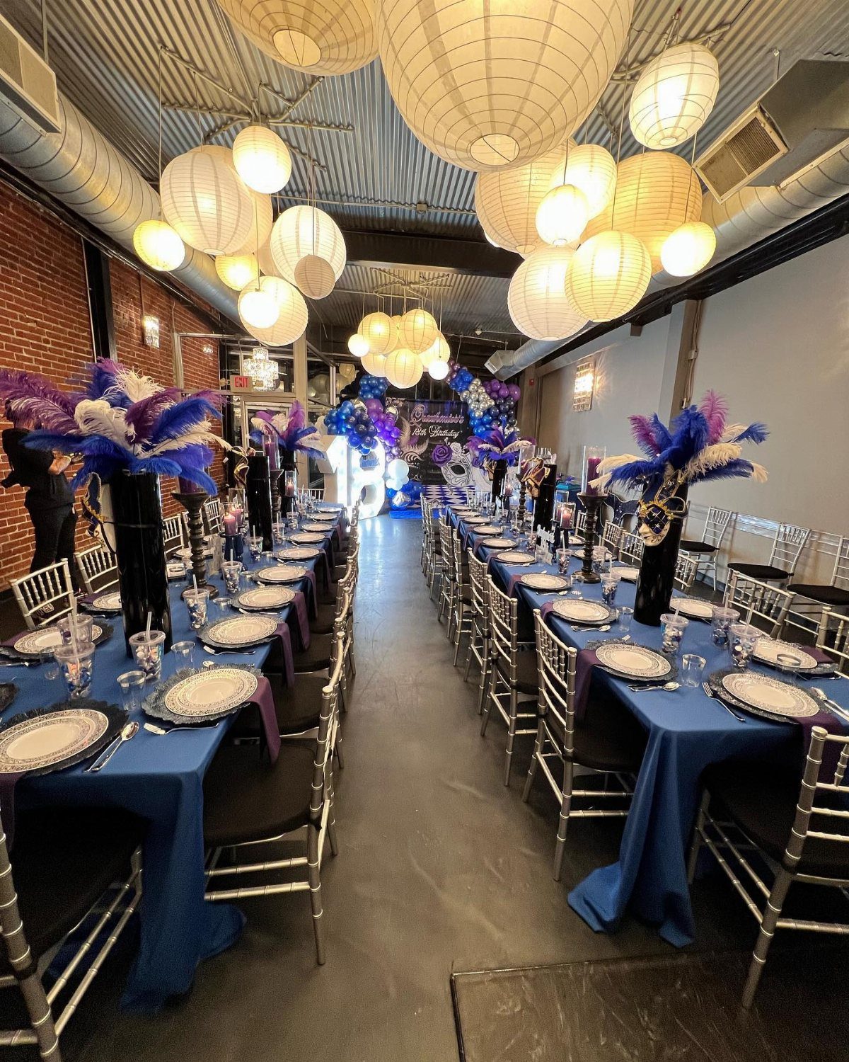 Two long banquet tables with blue cloths and feather centerpieces under hanging white paper lanterns.