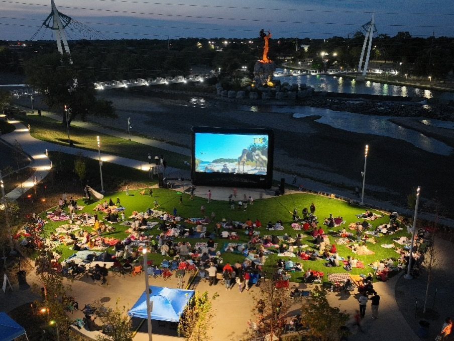 People sitting on blankets on a lawn watching a movie on a large outdoor screen.