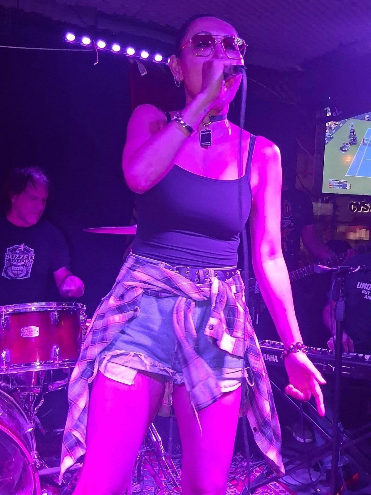 A woman singing into a microphone on a stage with purple lighting and a drummer.