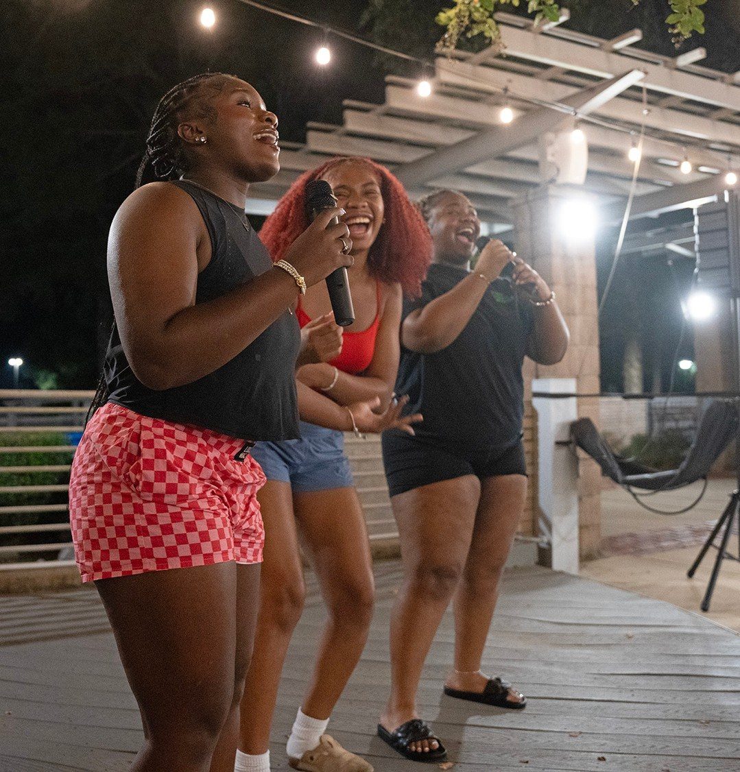 Three young women laughing and singing into microphones during an outdoor night party.