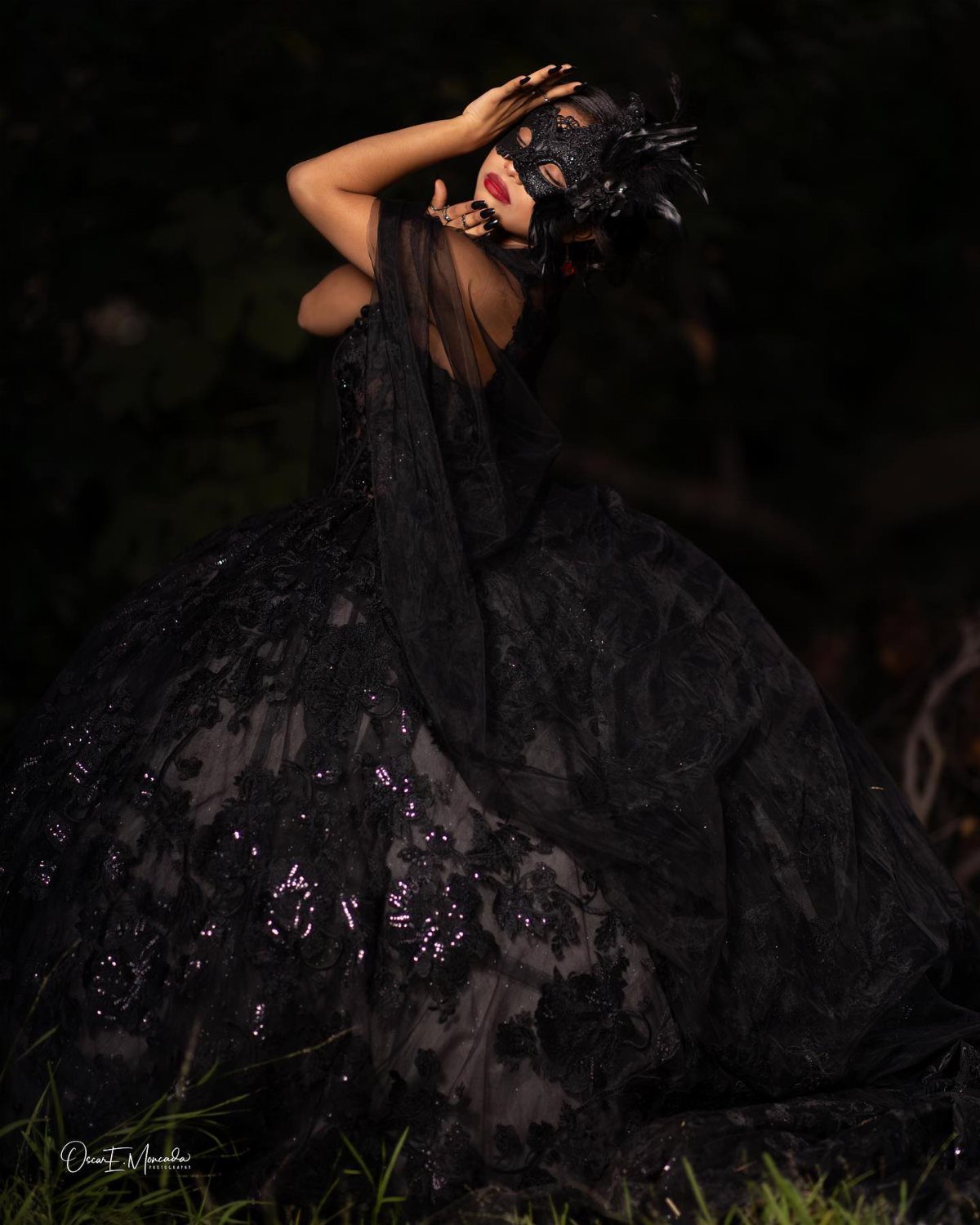 A person in a black lace gown and feathered masquerade mask posing outdoors.