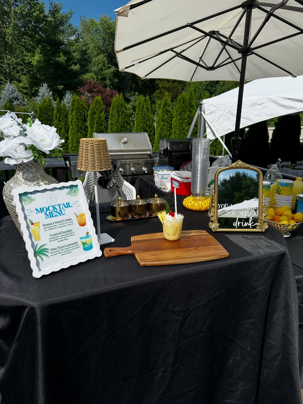 A festive mocktail bar setup with a menu, garnished drinks, and white flowers outdoors.