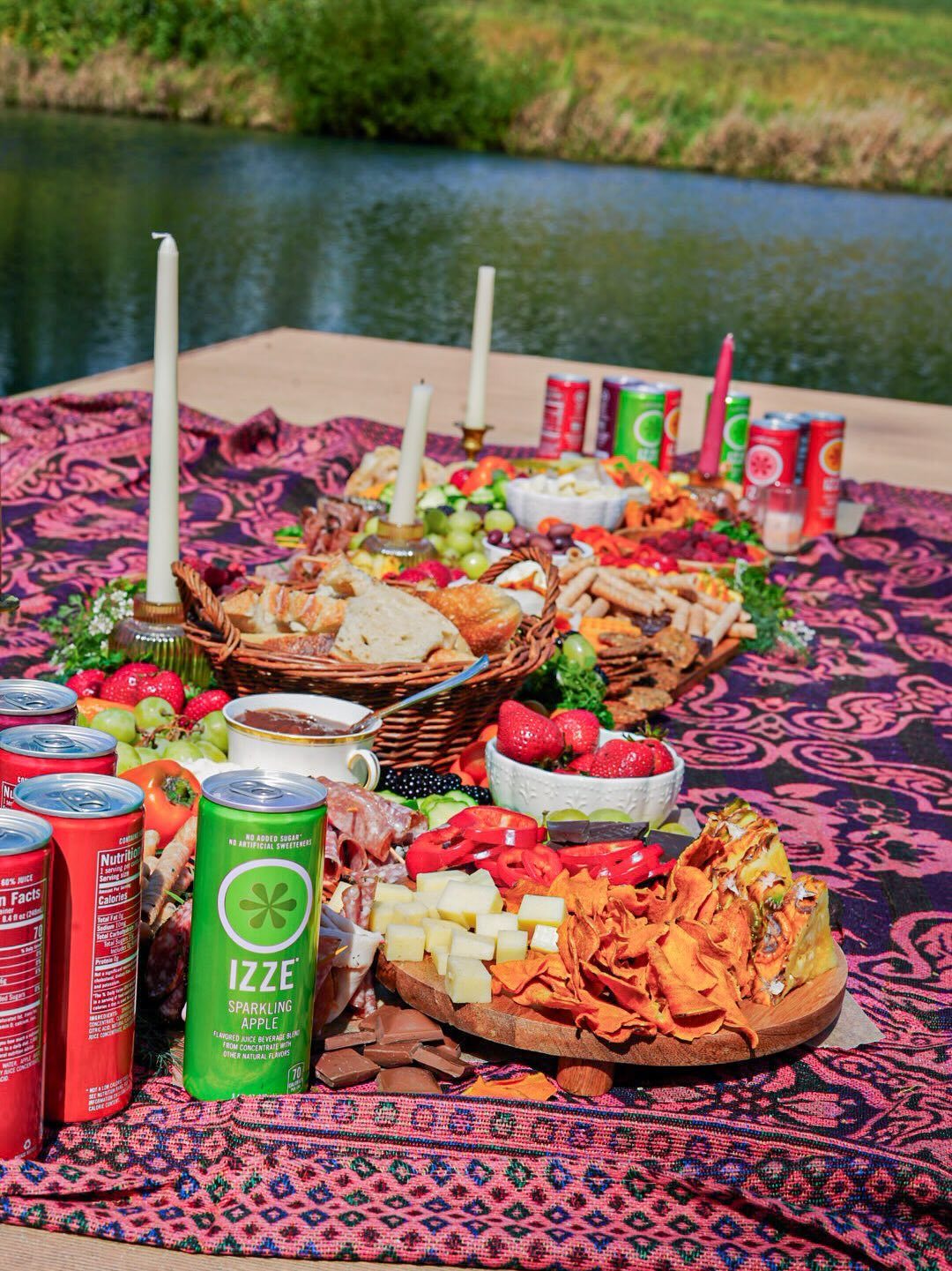 A large outdoor picnic spread with bread, cheese, fruit, and sparkling juice by a river.
