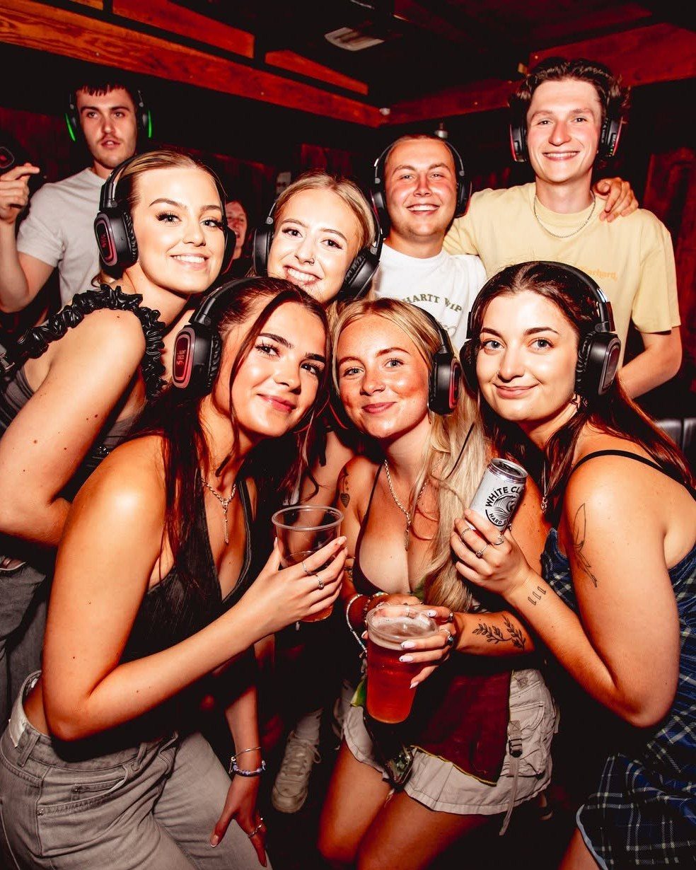 Six young adults wearing headphones and smiling together during a night party.