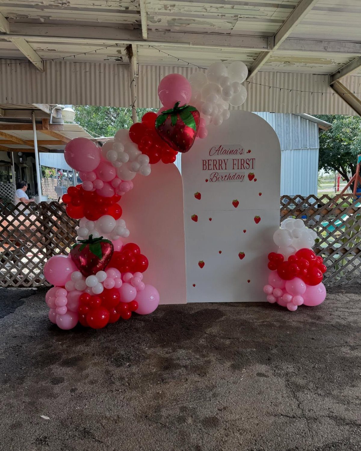 White arched birthday backdrop decorated with red, pink, and white balloon garlands and strawberry foil balloons.