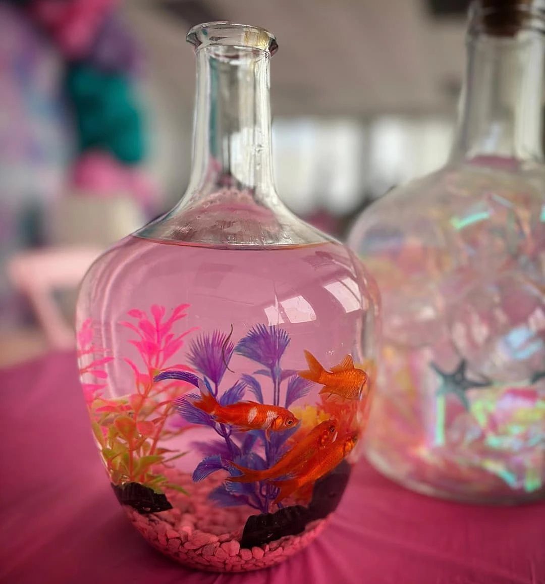 Glass bottle centerpiece containing pink liquid, colorful artificial fish, and decorative aquatic plants.