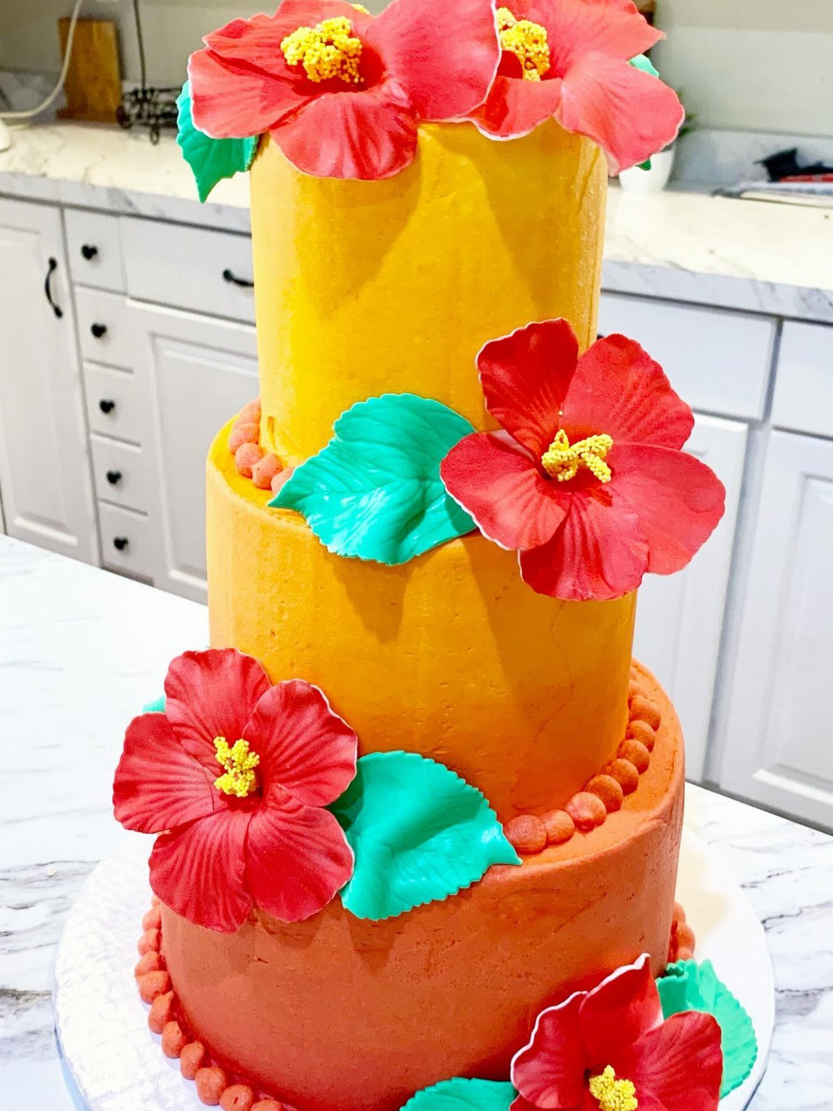 Three-tiered orange and yellow cake decorated with large red hibiscus flowers and teal leaves.