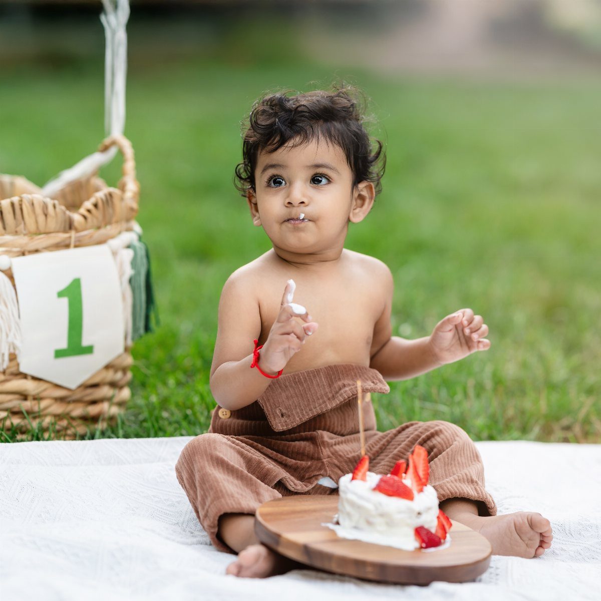 Toddler sitting on a white blanket outdoors with a small strawberry cake in front.