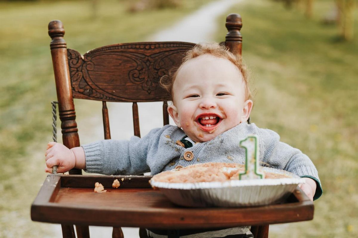 Smiling toddler with food on face sitting in a high chair with a pie.
