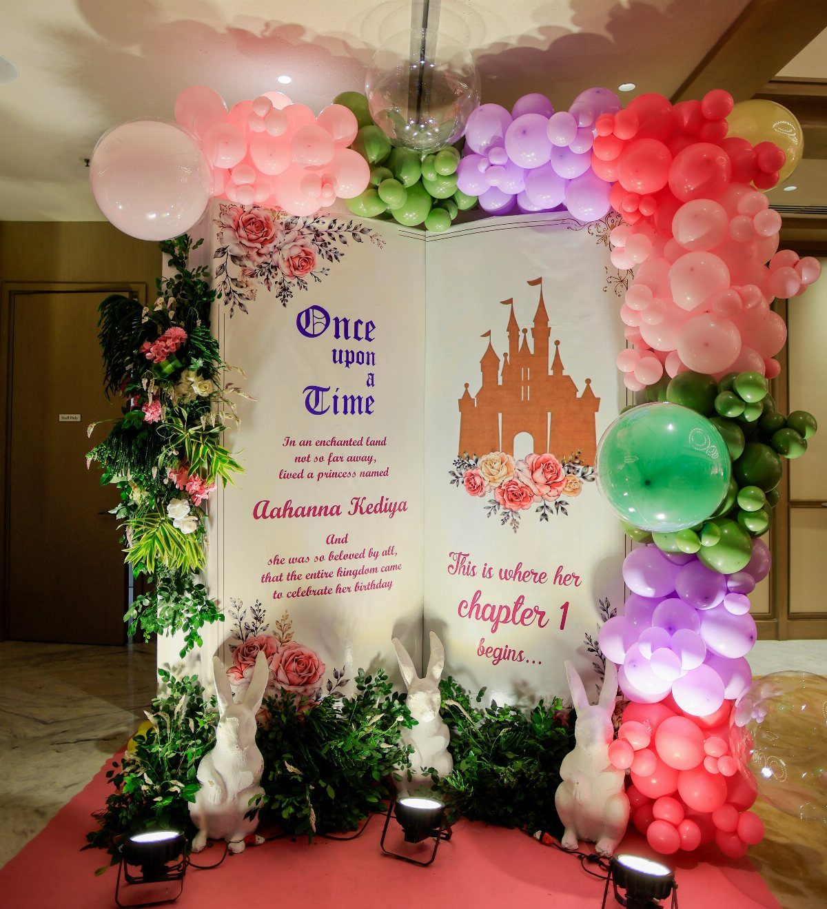 A fairy tale themed birthday backdrop featuring an open book, colorful balloon arches, and white rabbit statues.
