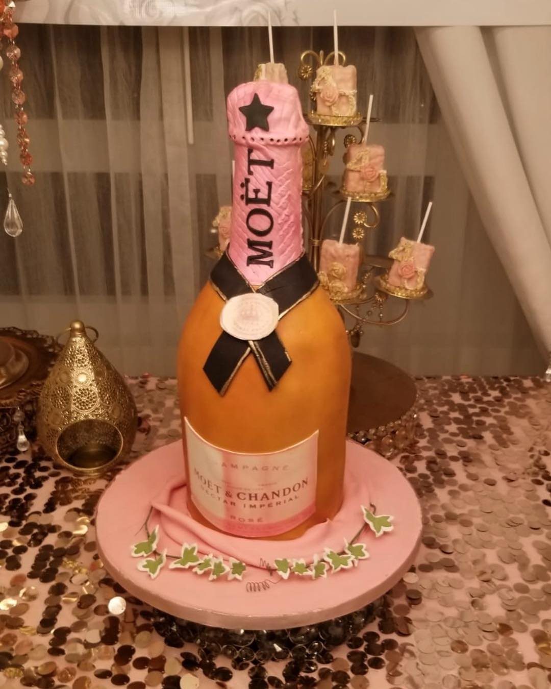 3D champagne bottle cake with gold and pink design on a decorative table.