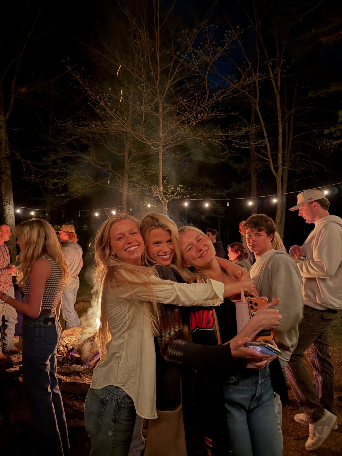 Three friends hugging at a bonfire party in a forest.
