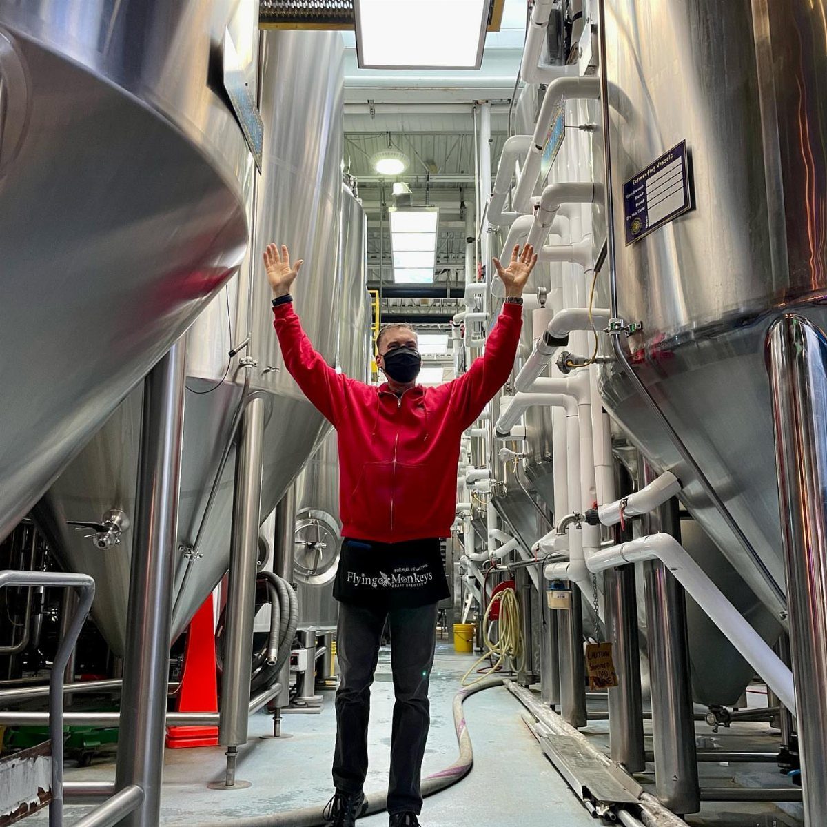 Man in a red jacket raising his arms between large stainless steel brewery tanks.