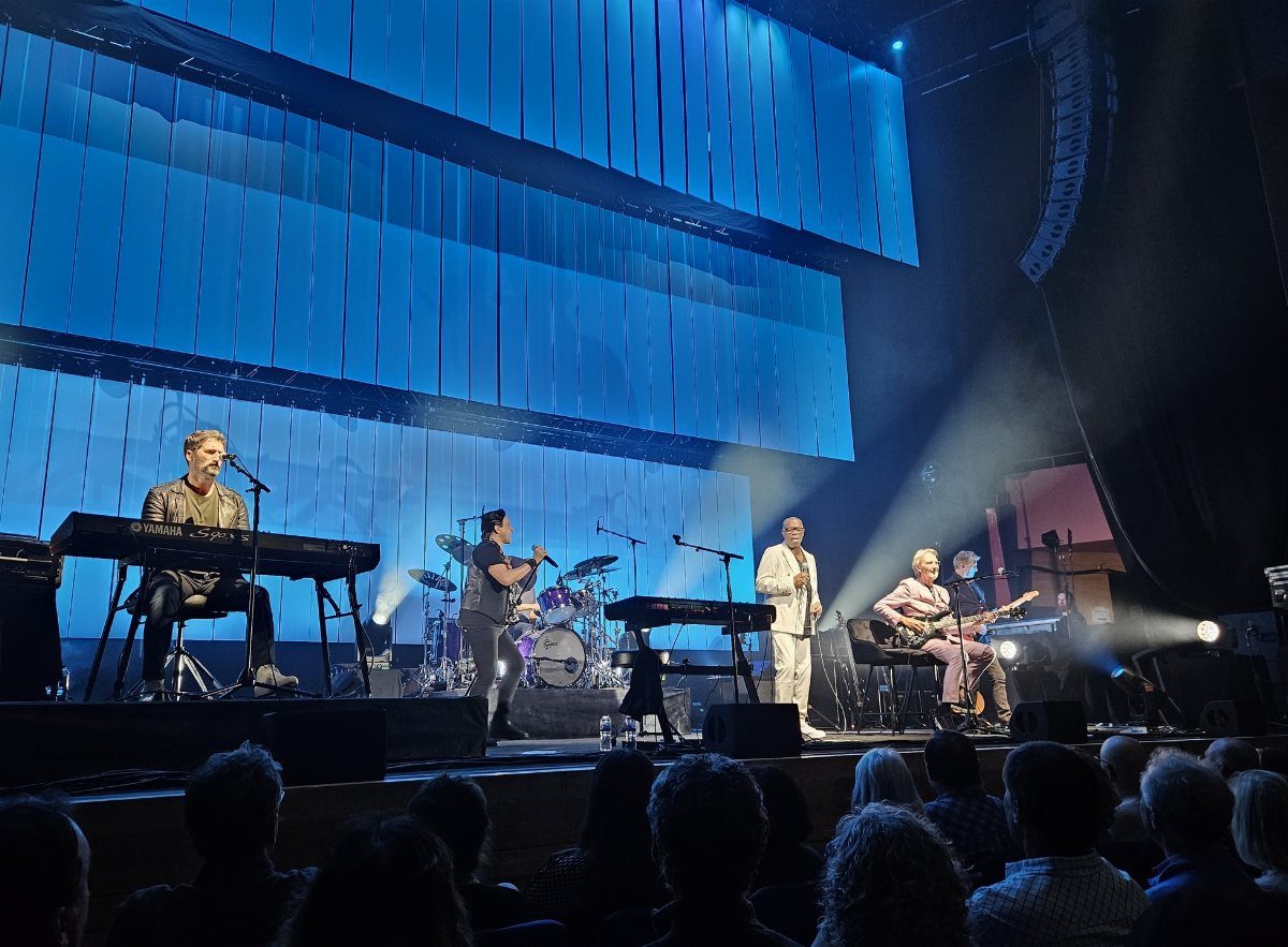 Band performing on stage with blue lighting and audience in foreground.