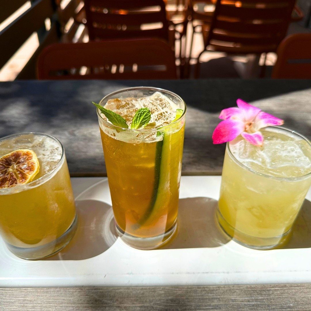 Three colorful iced cocktails with fruit and flower garnishes on a white tray.