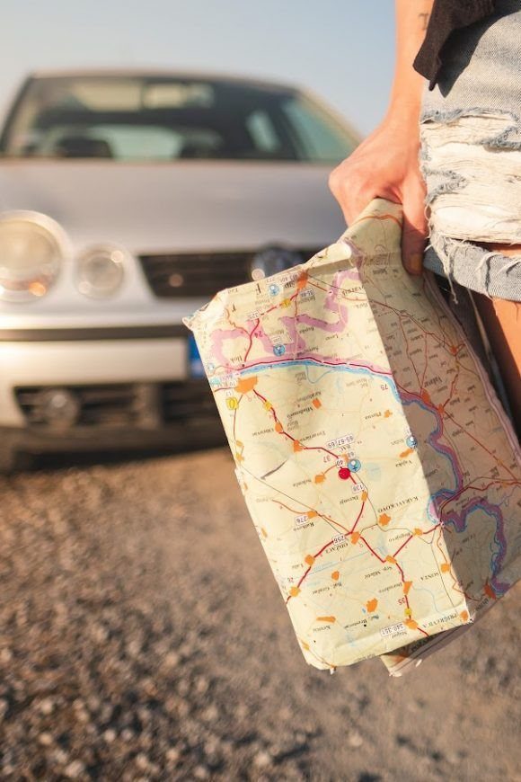 Person holding a road map near a parked silver car.
