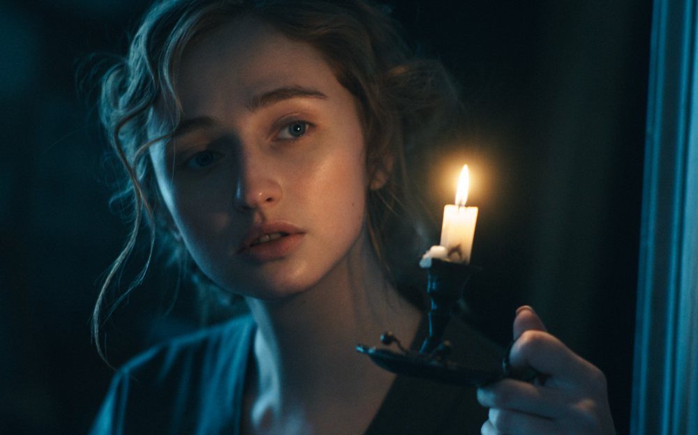 A young woman gazing pensively at a single lit candle held in a dark room.