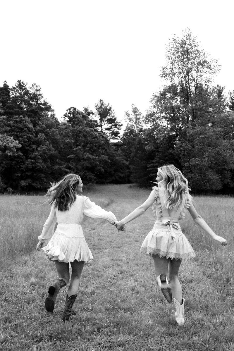 Two young women in dresses running through a grassy field while holding hands.