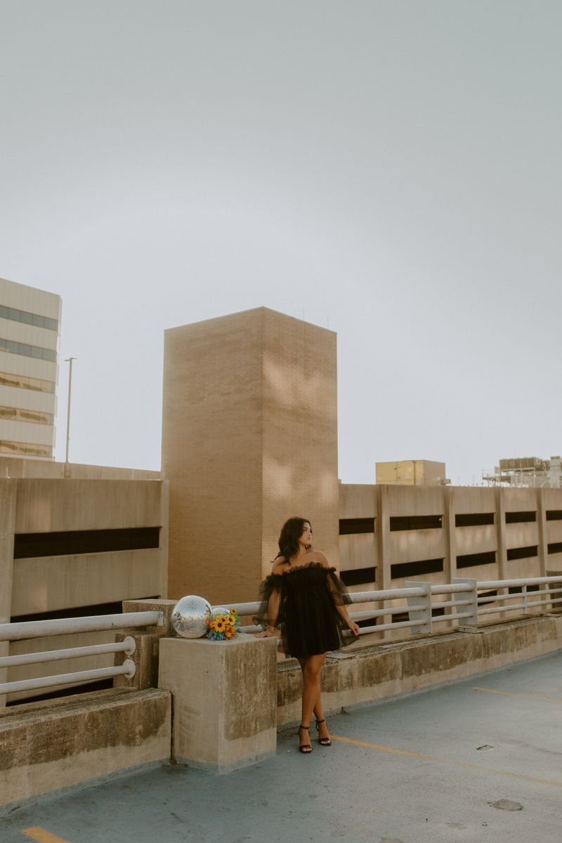 A woman in a black dress poses on a rooftop with city skyline and colorful balloons.