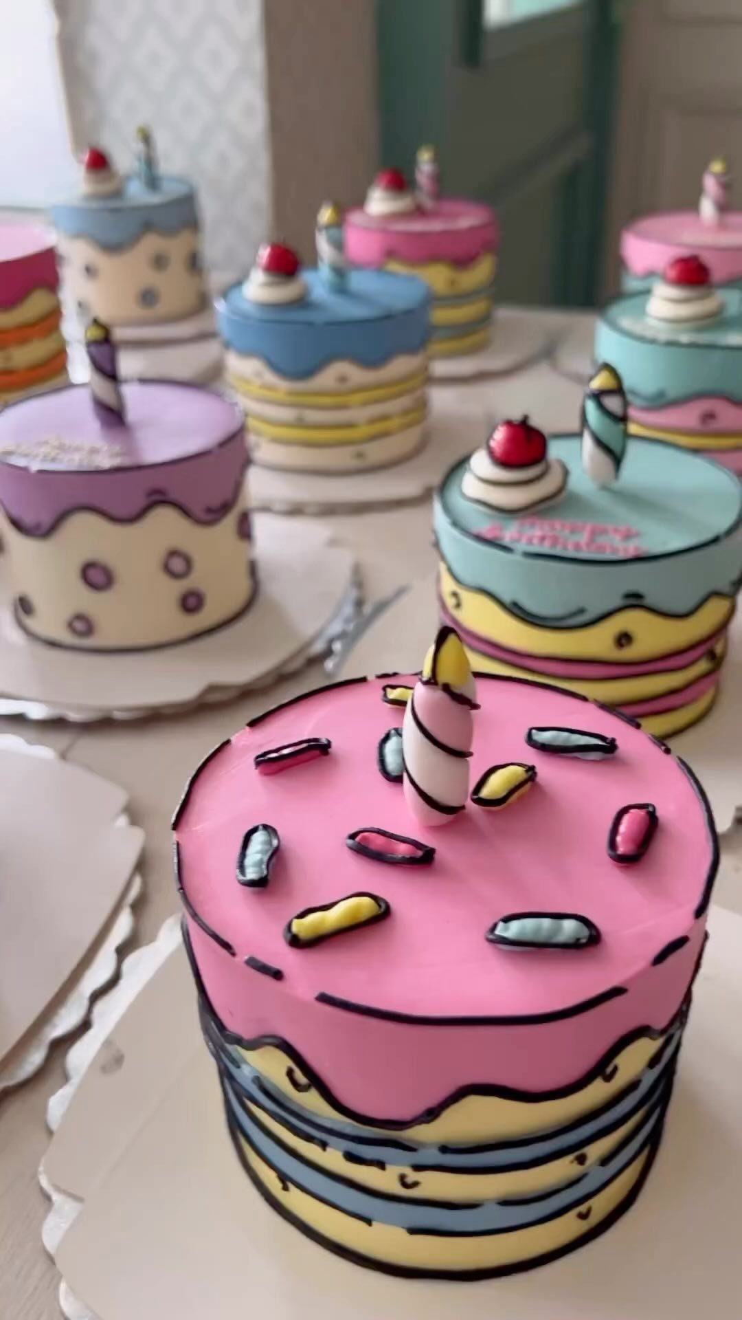 Multiple small pastel cakes with thick black outlines and single candles on white bases.