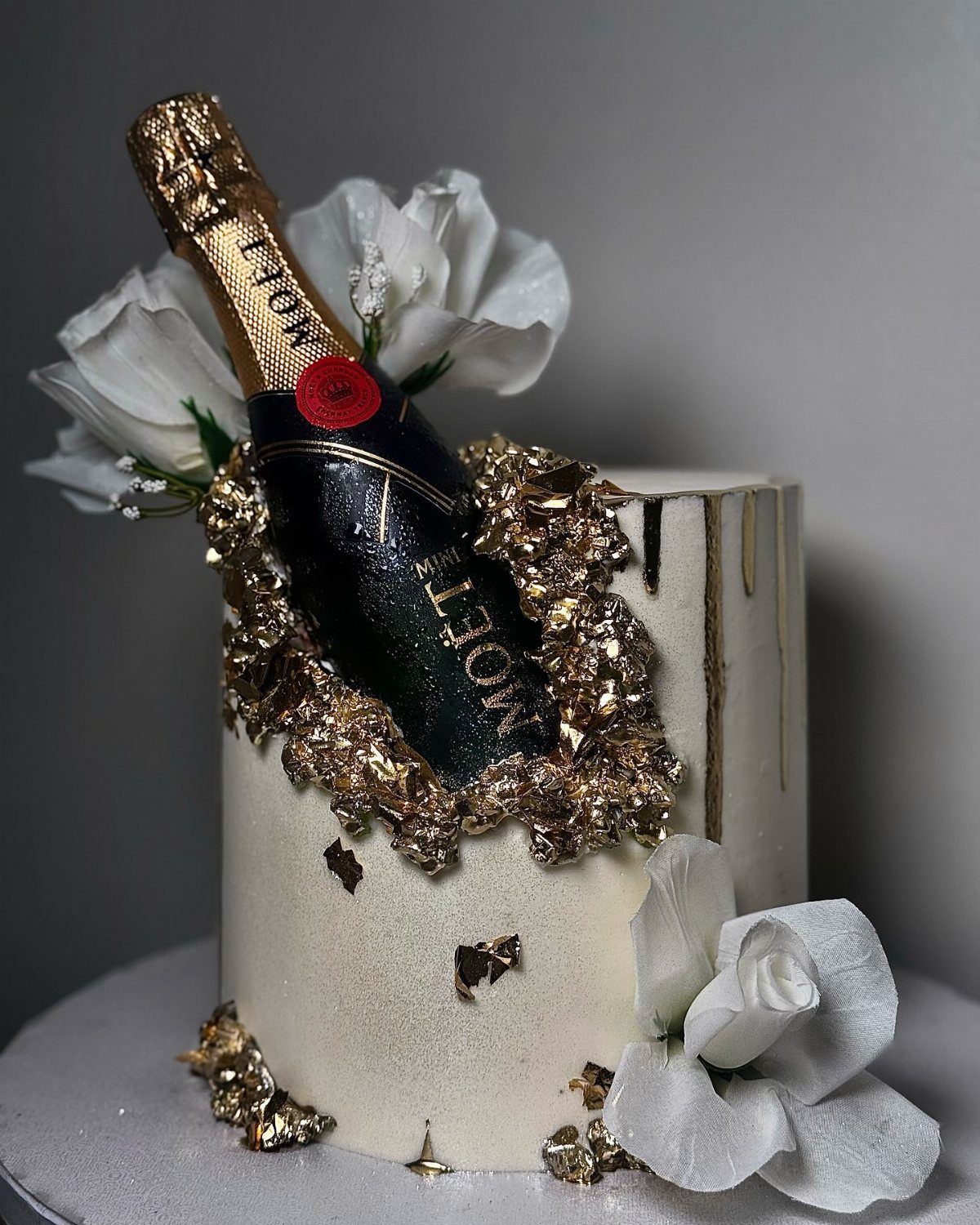 White cake decorated with gold leaf, white flowers, and a miniature Moët champagne bottle.