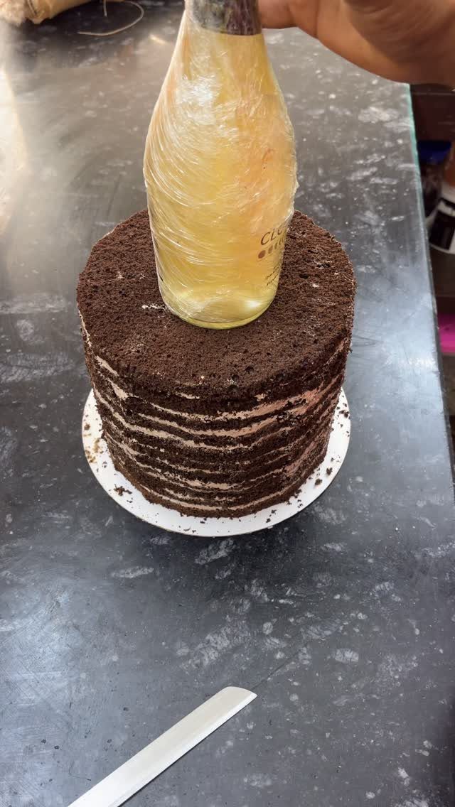 A hand pushing a champagne bottle into a layered chocolate cake on a dark counter.