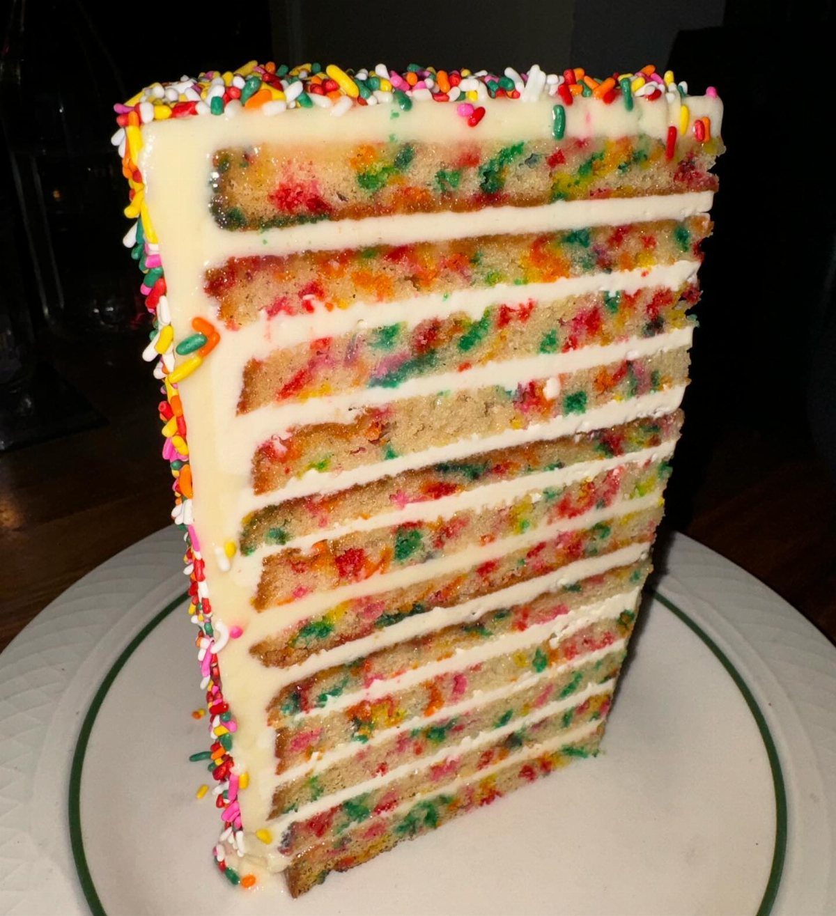A tall slice of multi-layered funfetti cake with white frosting and rainbow sprinkles on a plate.