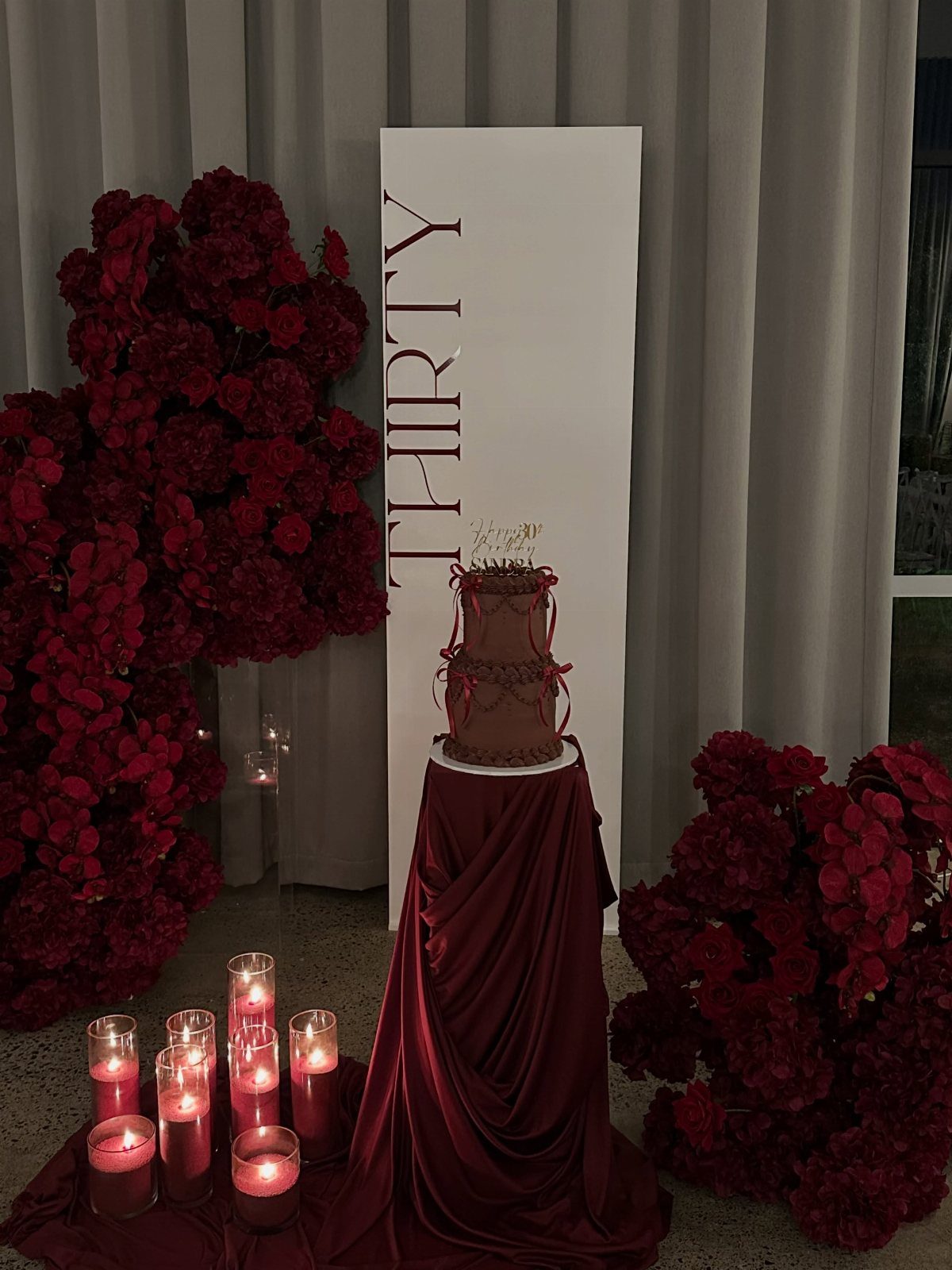A dark red tiered cake with bows stands among red flowers and lit candles.