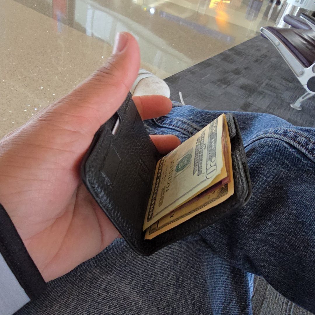 A hand holding open a slim black leather wallet filled with twenty-dollar bills.